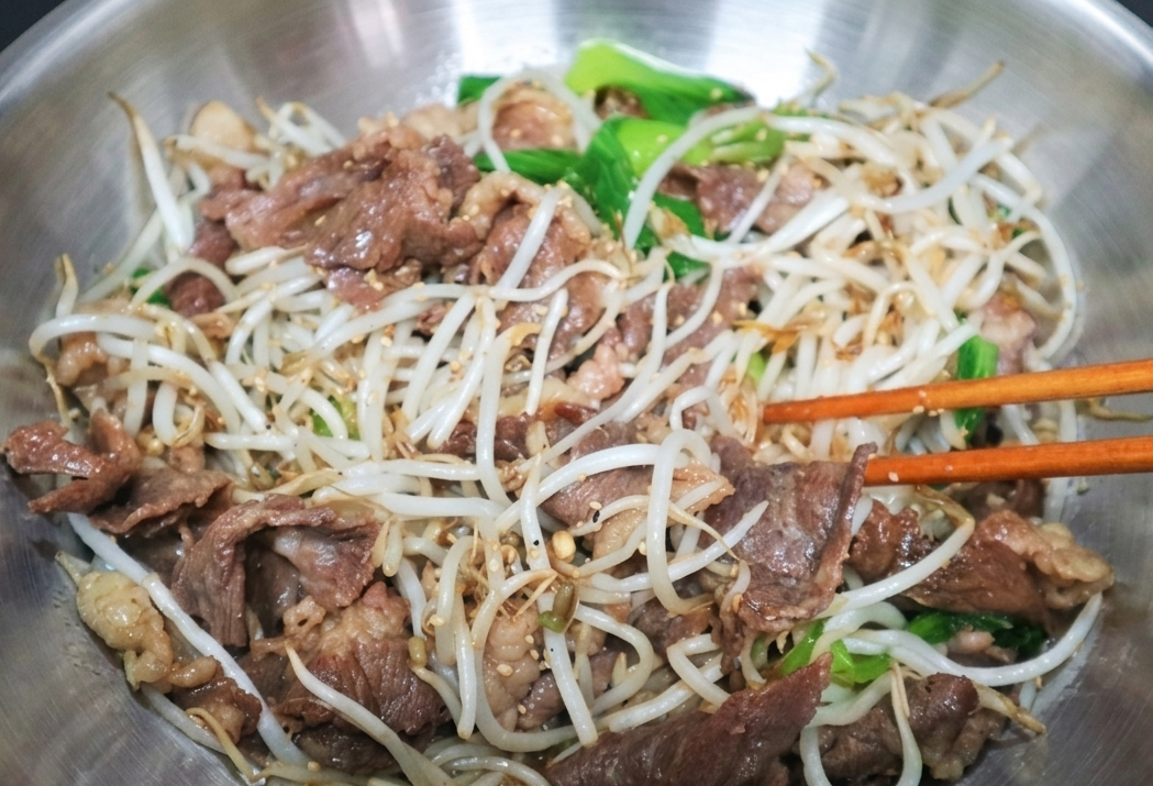 차돌박이 소고기와 싱싱한 숙주, 초록빛 야채가 양념과 함께 어우러져 볶아지고 있는 모습. 나무 젓가락으로 재료를 고루 섞으며 조리하는 생동감 있는 주방의 풍경.