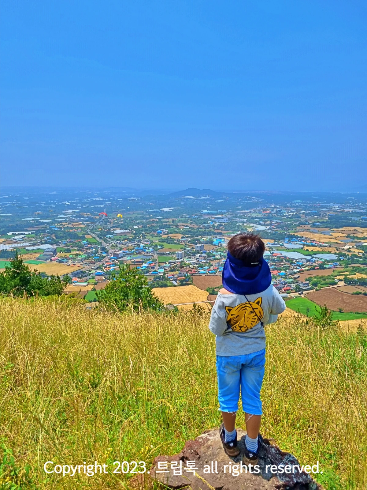 파란 하늘과 산 노란색 노과 밭, 도로 등의 마을이 애무 작에 보이는 금오름 정상에 서있는 아이의 뒷모습