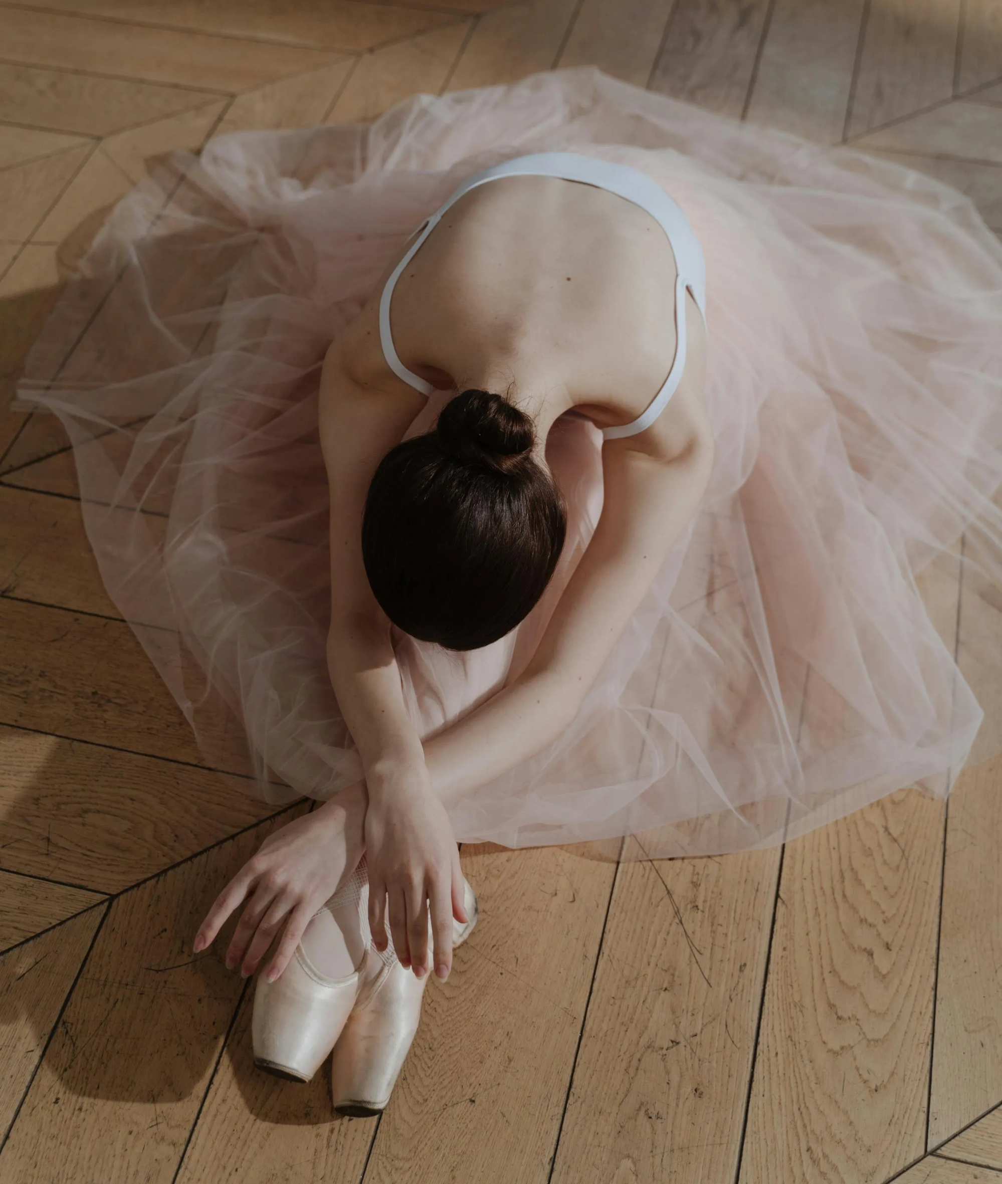 연한 핑크 튀튀를 입은 발레리나가 연습실 바닥 위에서 상체를 숙이며 스트레칭하는 모습