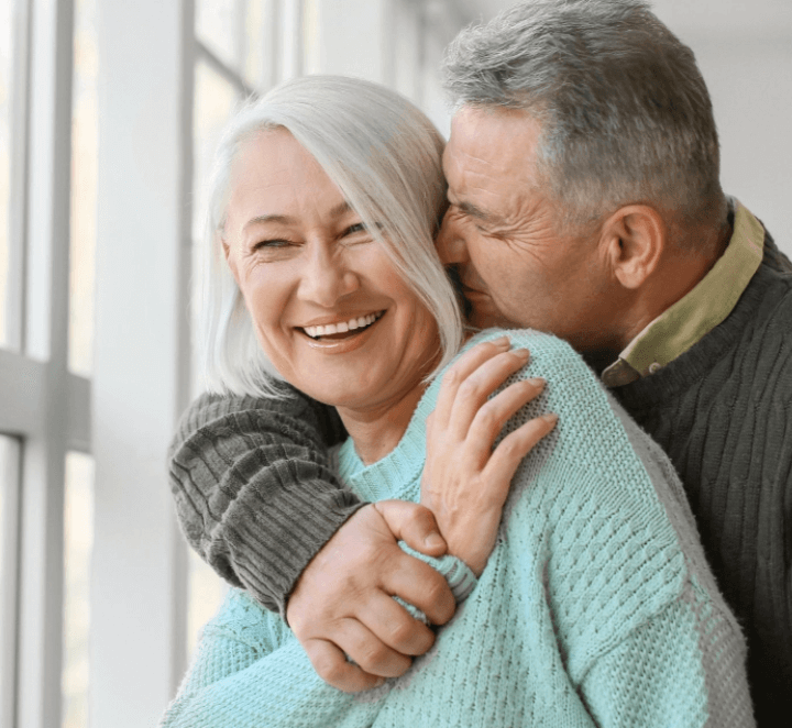 서양의 머리 희끗한 노부부가 백허그하며 다정히 웃는 모습