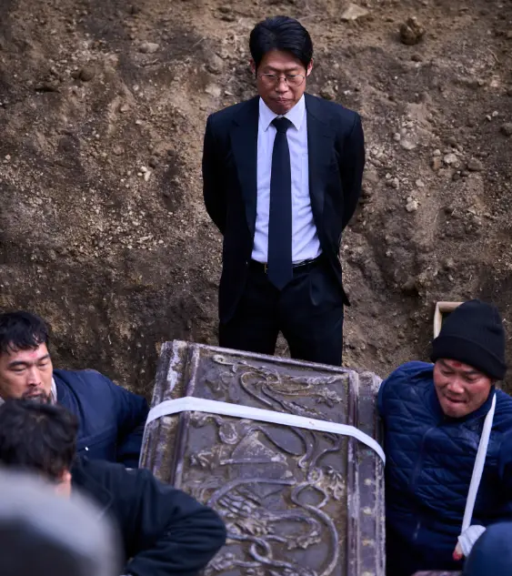 파묘- 검정색 흙 가운데 파란옷을 입은 일꾼들이 묘를 들고 나가고 양복에 와이셔츠 넥타이를 한 유해진이 진지하게 이를 바라보고 있는 모습