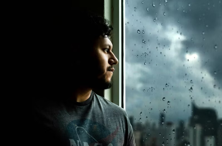 창문 너머 비 내리는 풍경을 바라보며 실내에 앉아 있는 사람. 흐린 날씨와 함께 고요한 분위기 속에서 사색하는 모습이 담겨 있음.