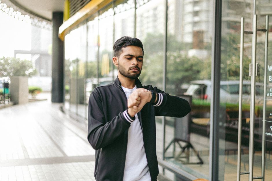 A man in a city looks at his wristwatch while standing near a modern glass building.