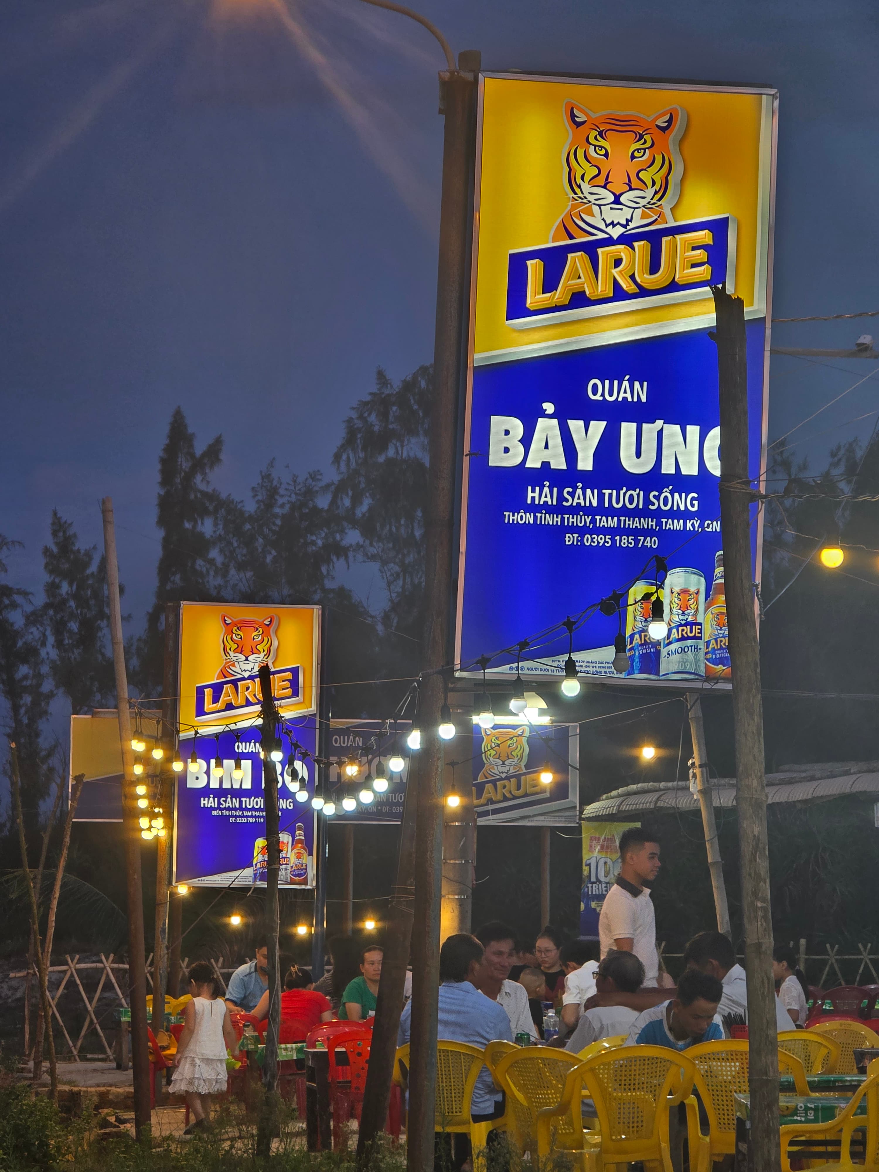 베트남 다낭 땀끼 해변의 해산물 음식점 야경. 노란&middot;빨간 플라스틱 의자에 앉아 많은 사람들이 식사를 즐기고 있고, 머리 위로 전구 조명이 반짝인다. 간판에는 'Larue' 맥주 광고와 'Bảy Ứng' 해산물 식당 이름이 보인다.