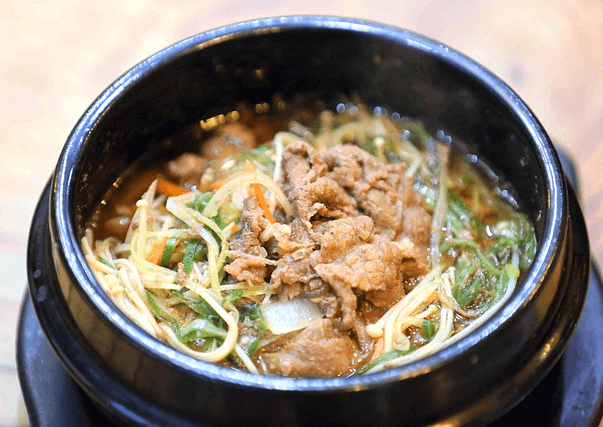 검정 뚝배기에 불고기백반이 가득하다. 익힌 돼지고기와 대파, 야채, 국물이 있다.