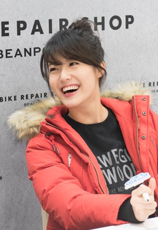 'REPAIR SHOP' 글자가 보이는 배경 앞에서 빨간 패딩 점퍼를 입고 웃는 짧은 머리의 여성