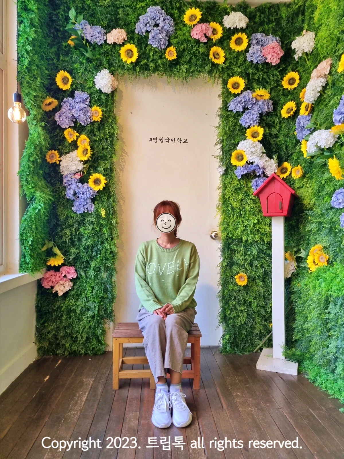 벽을 초록덩쿨과 노란 해바라기 보랗고 분홍색 꽃으로 장식되고 가운에 의자에 한 사람이 앉아있다.