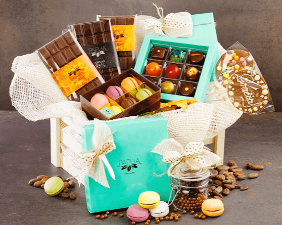 나무 박스에 잘 포장된 초콜릿 선물들이 가득하다. 청록색 상자에 담긴 알록달록한 초콜릿들과 네모난 초콜릿들도 풍성하다. 주변에는 마카롱도 있다.