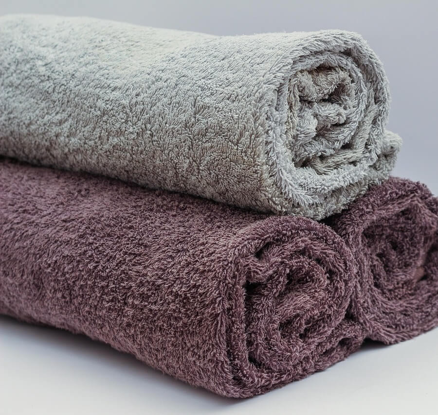 흰색 배경 앞에 연회색과 보라색 수건 세 장이 호텔식으로 돌돌 말려 깔끔하게 쌓여 있는 모습