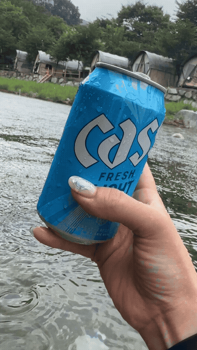 계곡 한가운데에서 손에 든 카스 라이트 맥주 캔. 물방울이 맺힌 맥주와 계곡 풍경이 시원함을 강조한다.