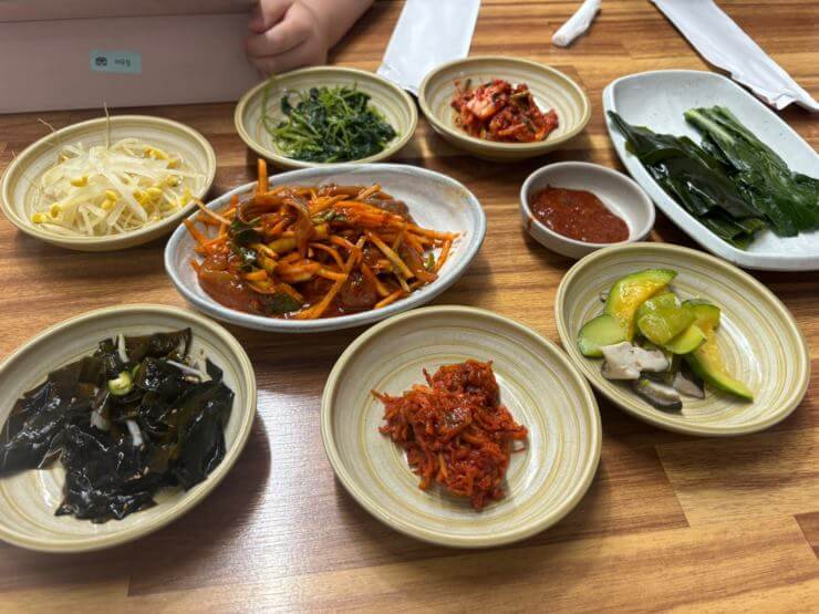 9개 반찬이 베이지색 접시에 담겨있다.
콩나물 미역무침 애호박볶음, 참나물무침, 김치가 동그랗게 놓여있다. 이 반찬들 가운데 하얀 접시에 빨갛게 무쳐진 멸치회무침이 있다.
오른쪽 위에는 긴 접시 위에 다시마와 데친 케일이 있다.