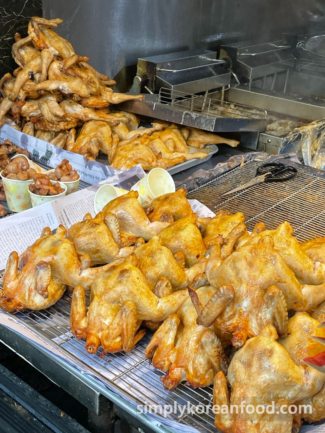 Dozens of crispy whole chickens cooling on wire racks at Korea Tongdak in Dongmyo, surrounded by chicken gizzards and deep fryers in the background.