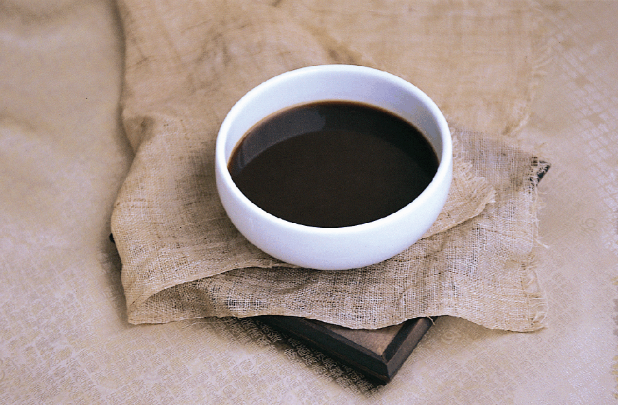 한약재를 물과 함께 넣고 달여서 만든 검은 색의 한약을 희고 동그란 그릇에 반쯤 담아둔 것을 황금색 바닥 위에 검고 네모난 받침과 그 위에 삼베를 놓아둔 곳에 두고 찍은 사진