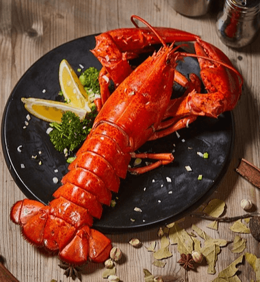 동그란 검정 접시에 붉은 랍스터 한 마리가 있고 왼쪽에는 노란 레몬 조각들과 파슬리가 있다.