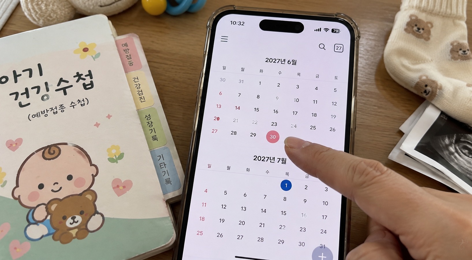 아기 건강수첩과 양말 옆에서 스마트폰 달력으로 2027년 6월 30일과 7월 1일 출생일을 비교하는 모습