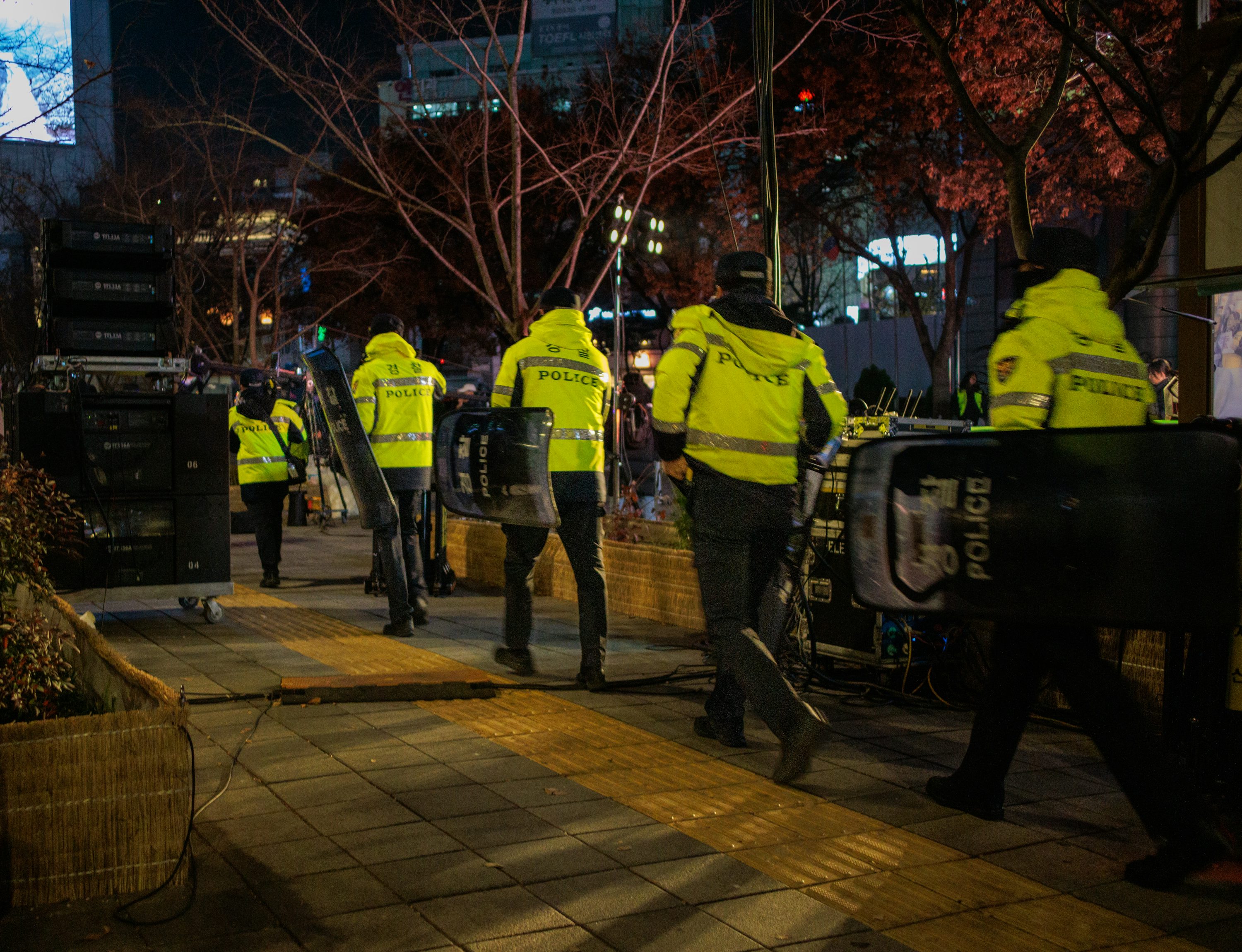 야간에 형광 노란색 POLICE 조끼를 입은 한국 경찰관들이 방패를 들고 이동하는 뒷모습