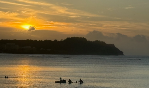 괌, 투몬 비치 해변의 해가 지는 광경. 하늘이 붉게 물들었고 바다에는 사람들이 일몰을 바라보고 있다.