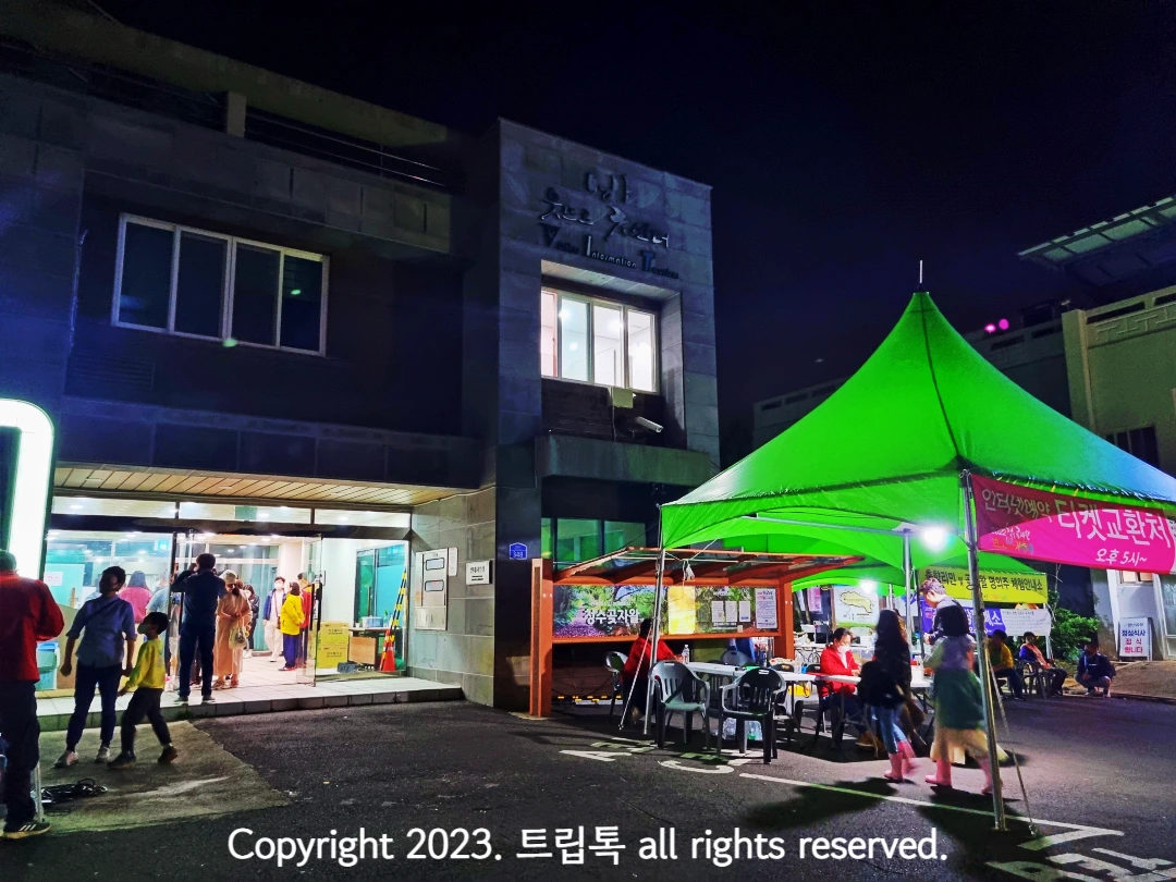왼쪽에 건물이 있고 로비에 사람이 많고 그 오른쪽에 연두색 천막이 있다.