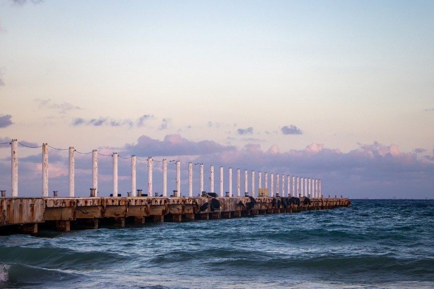 멕시코 플라야 델 카르멘 해변에 위치한 긴 목재 부두와 잔잔하게 일렁이는 카리브해 파도. 노을진 하늘 아래 평화로운 휴양지 풍경.