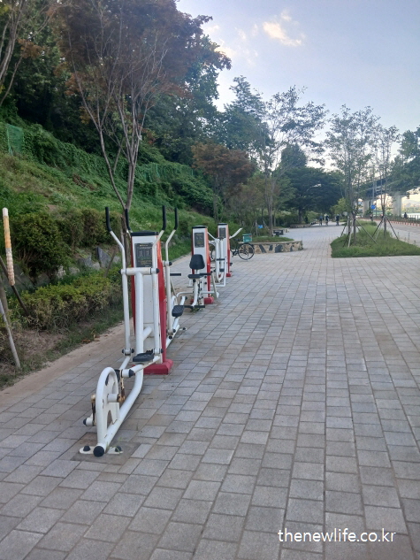 산책로 옆 공원에 설치된 하체 운동 중심의 흰색·빨간색 실외 운동기구 – 뱃살 빼는 운동 기초체력 단련용