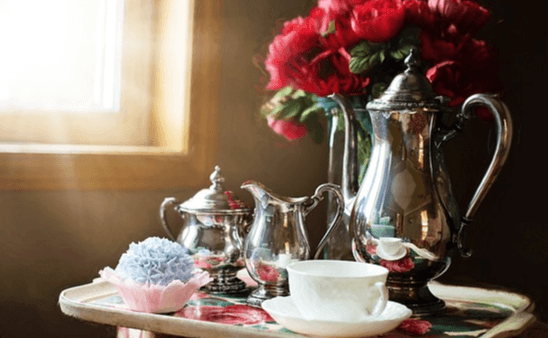 작은 탁자 위에 은색 주전자와 하얀 찻잔 세트가 있고 빨간 장미가 놓여있다.