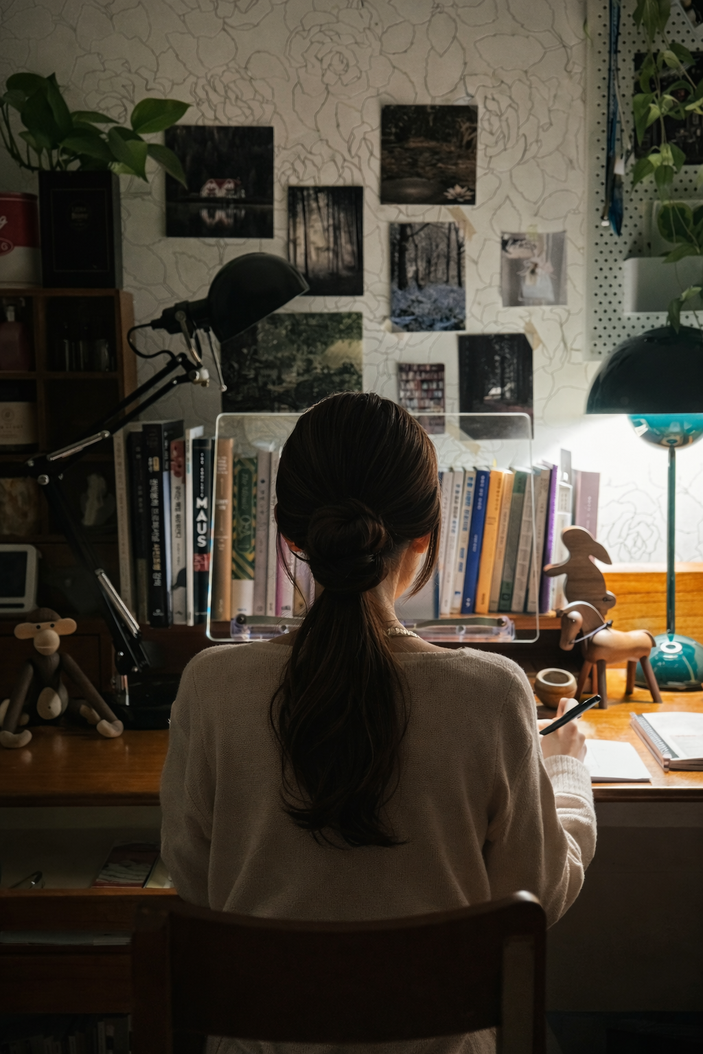 책으로 가득한 서재 책상 앞에 앉아 긴 머리를 우아하게 묶은 여성이 조용히 글을 쓰고 있는 뒷모습