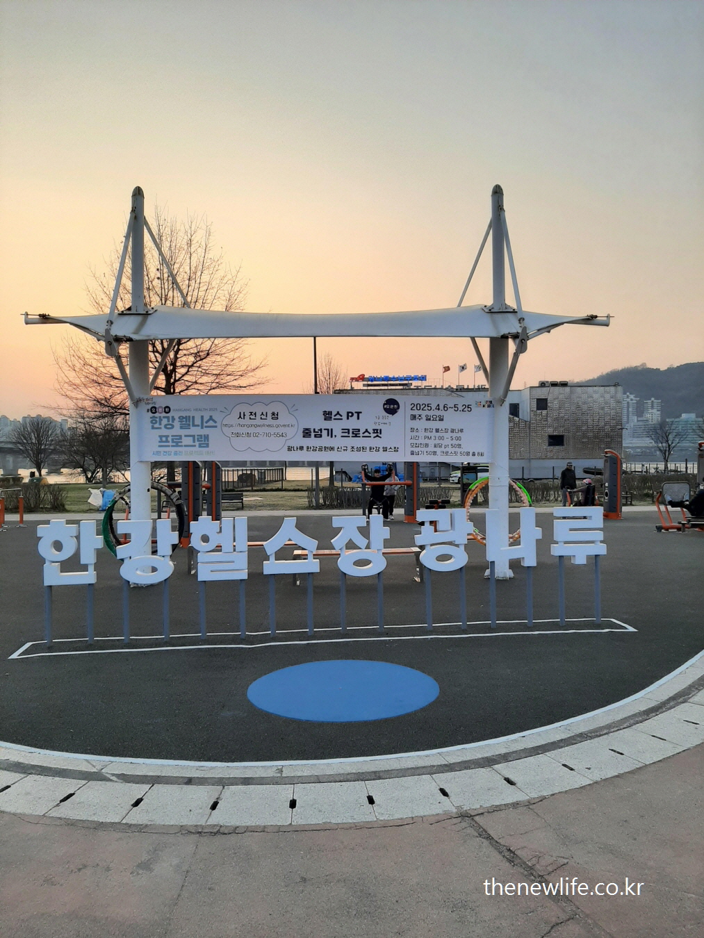 Outdoor fitness equipment and wellness program banner at Hangang Kwangnaru Health Park during sunset-광나루 한강공원 헬스장 전경. 한강 웰니스 프로그램 현수막과 야외 운동 기구가 설치된 모습.
