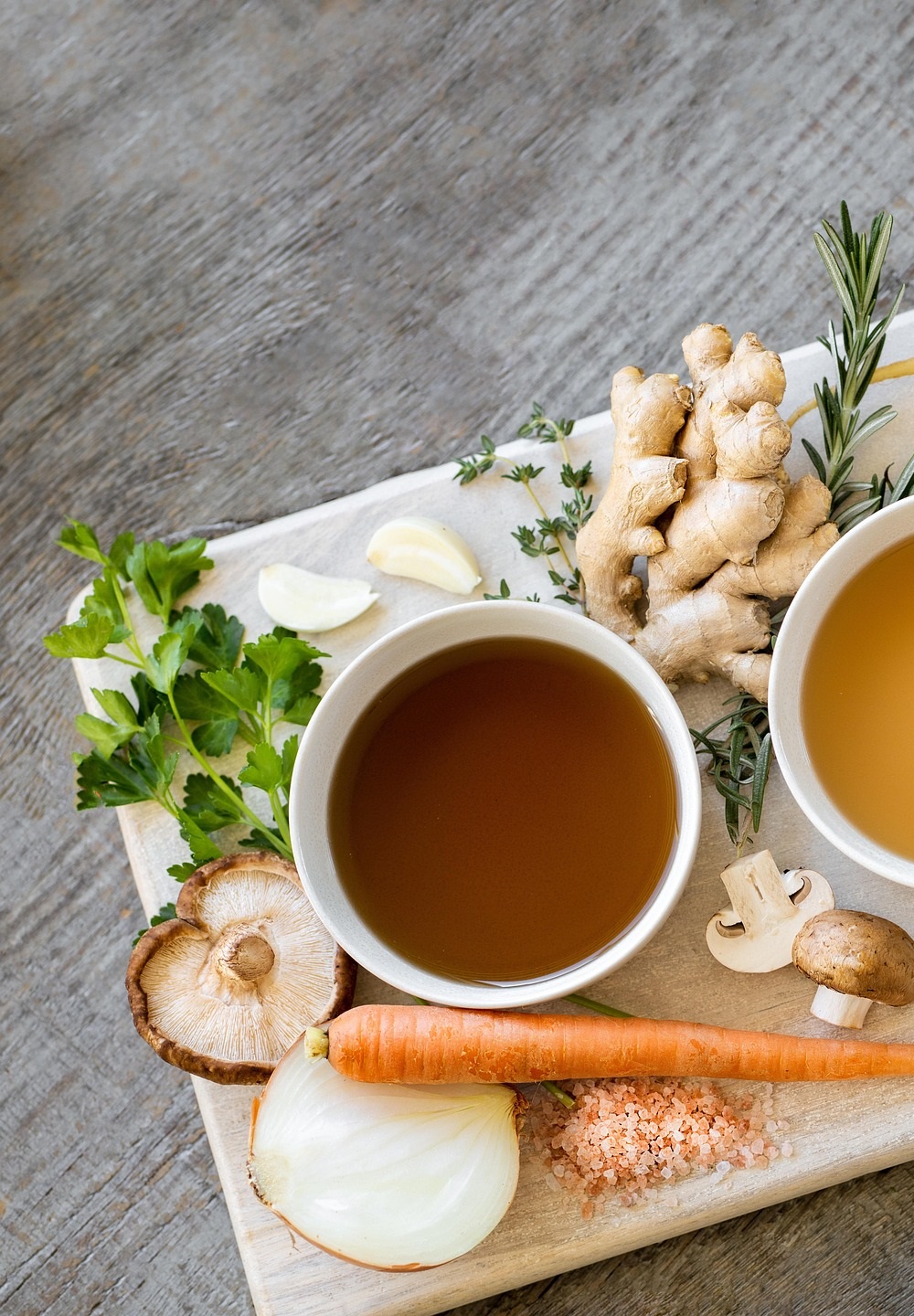 흰 그릇에 담긴 세 종류의 맑은 육수와 주변에 놓인 생강, 당근, 버섯, 마늘, 셀러리 등 신선한 식재료들