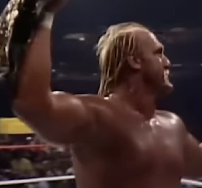 헐크 호건이 레슬링 경기장에서 챔피언 벨트를 높이 들어 올리며 승리를 자축하는 모습. 강인한 체격과 조명이 돋보인다.