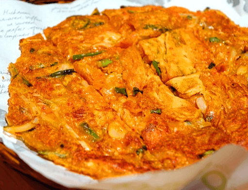 종이 위에 김치전 한 장이 놓여있다. 빨간 김치색을 가졌고 김치와 파 등이 보인다.