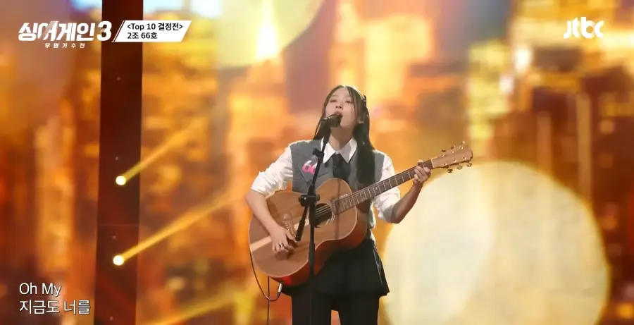 싱어게인3- 검정색 미니스커트에 흰셔츠&amp;#44; 회색 조끼를 입고 검은색 넥타이를 맨체 갈색기타를 들고 긴생머리 양쪽으로 삔을 꽂은채 스탠딩마이크 앞에서 노래하는 66호