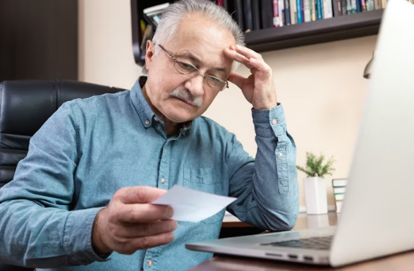건강보험 고지서를 들여다보며 걱정하는 중년 남성. 노트북 앞에 앉아 보험료 인상 내용을 확인하는 모습.