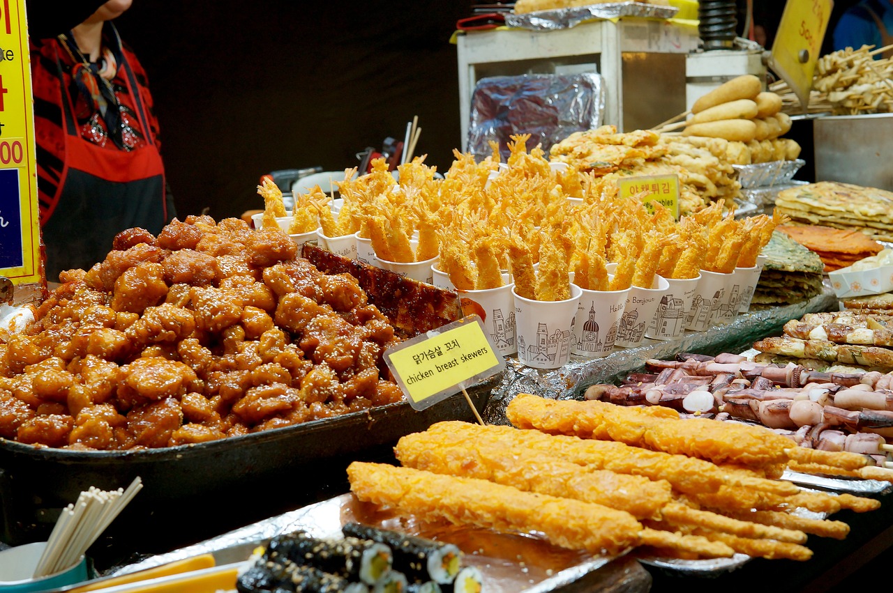 Korean street food stall selling sweet and spicy fried chicken, shrimp tempura in cups, skewers, and assorted snacks at a night market.