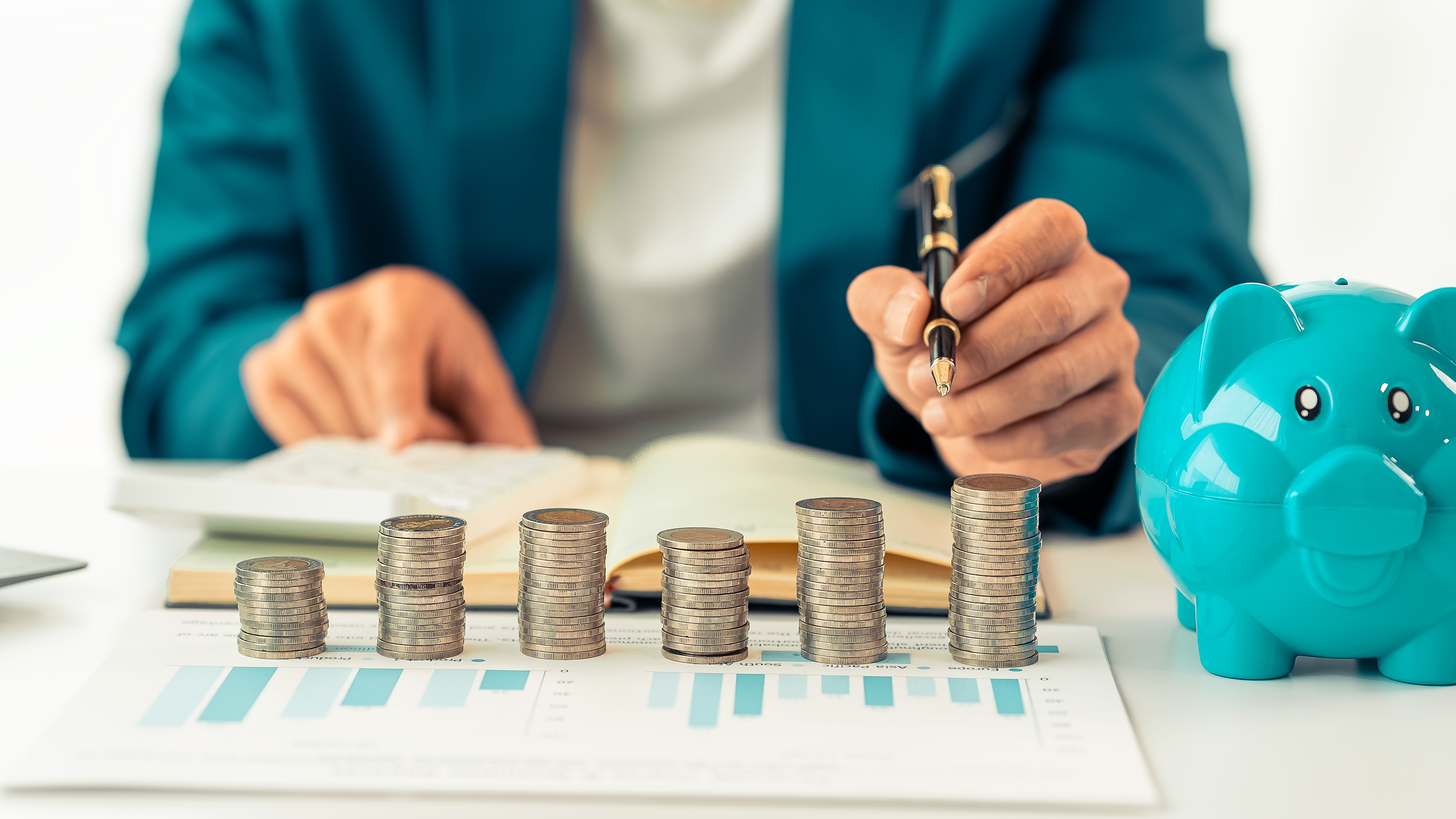 저축 및 자산관리를 분석하는 사람의 손과 쌓여 있는 동전들, 옆에는 파란 돼지 저금통이 있는 이미지. 청년 통장 제도 선택을 상징하는 장면