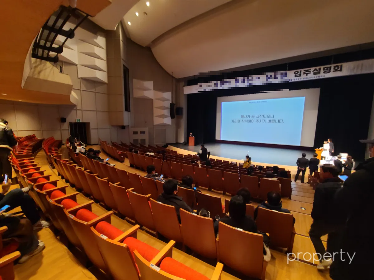 입주설명회장소-빨간색좌석-곳곳에앉은사람들-무대위에ppt화면이띄워져있고-무대상단에입주설명회현수막이달림