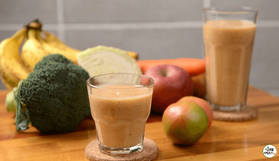 나무색식탁위에-바나나-브로콜리-양배추-사과-당근-토마토가-있고-과일과-야채를-갈아서-담은-유리컵이-2개-놓여져있다