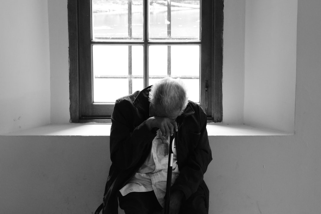 창가 의자에 홀로 앉아 고개를 숙이고 있는 노인의 흑백 사진