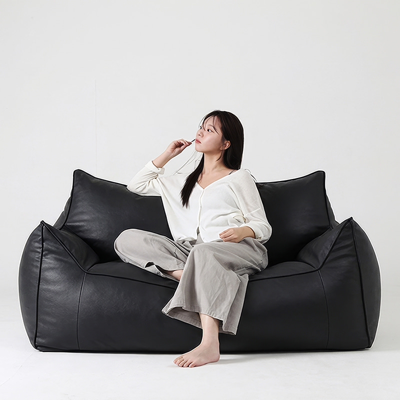 흰 배경에서 긴 머리 여성이 흰 가디건과 회색 바지 차림으로 맨발로 검은색 플래지어 톰빈백 2인용 기능성 가죽 빈백 소파에 앉아 한 손을 입가에 대고 옆을 바라보는 모습