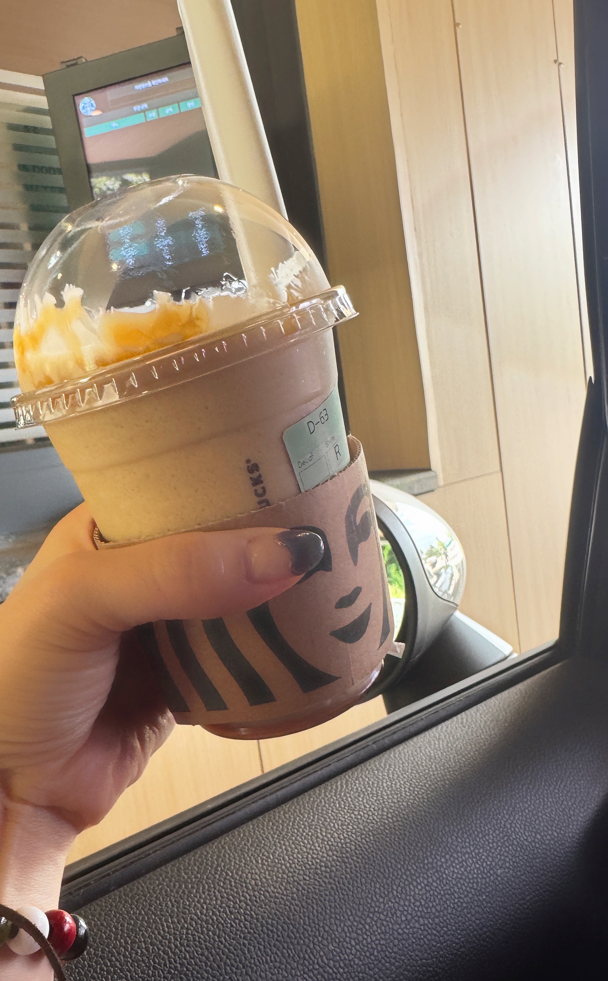 운전 중 드라이브 스루에서 구입한 스타벅스 프라푸치노를 손에 들고 있는 모습. 휘핑크림 위에 카라멜 시럽이 얹혀 있으며, 배경으로 매장 벽과 사이드미러가 보인다.