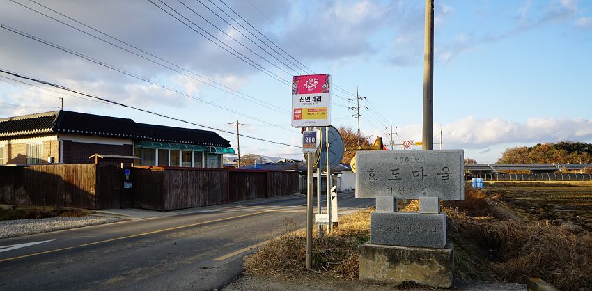 마을 앞 차로 변 회색 대리석에 새겨진 '효도마을 2001', 뒤에 검은 지붕의 단층 건물, 좌 우로 지나가는 전깃줄 여러줄, 푸른 하늘, 흰구름,