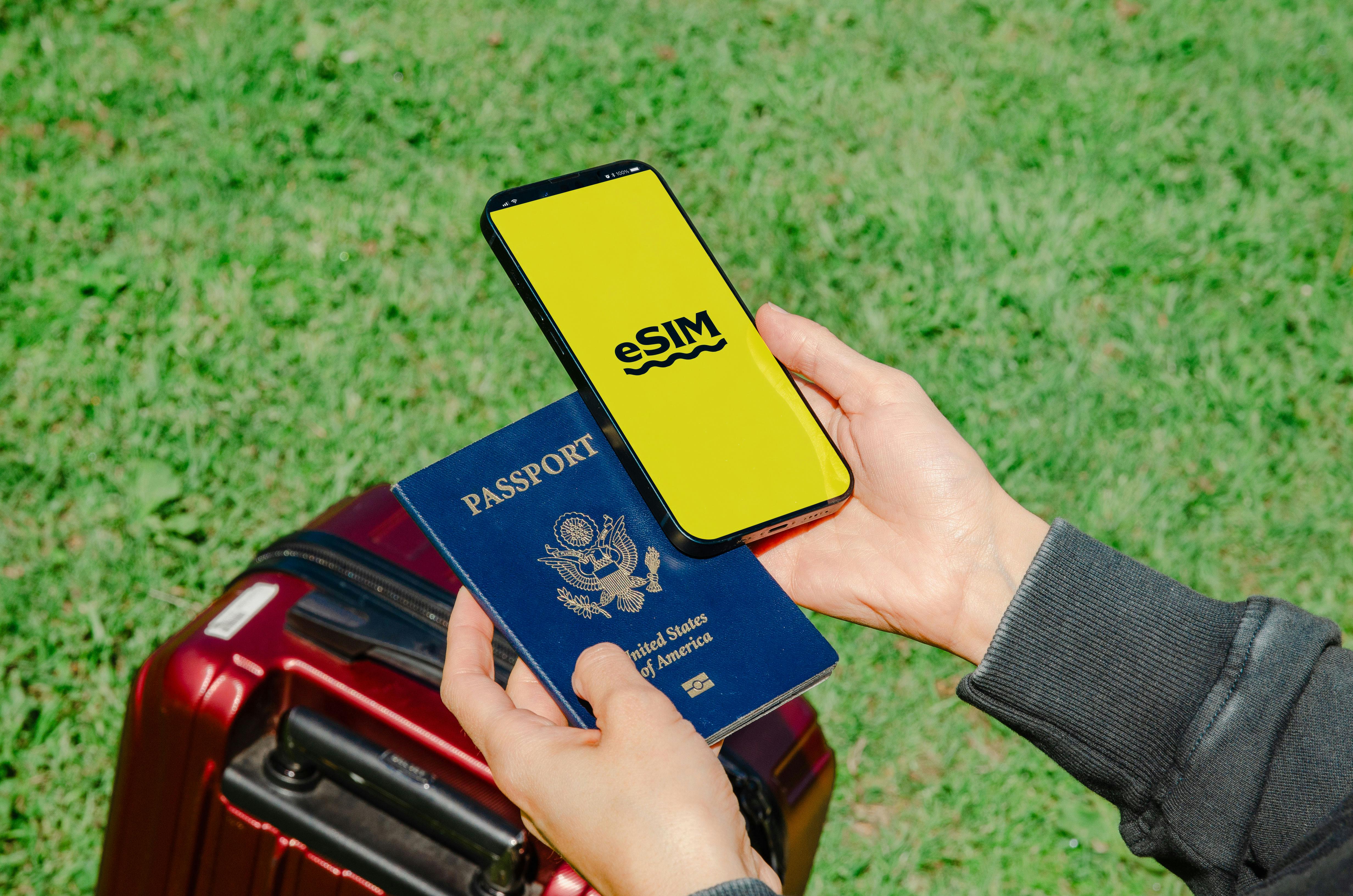 잔디밭에서 SIM 화면이 뜬 스마트폰과 여권을 든 손, 여행가방 일부