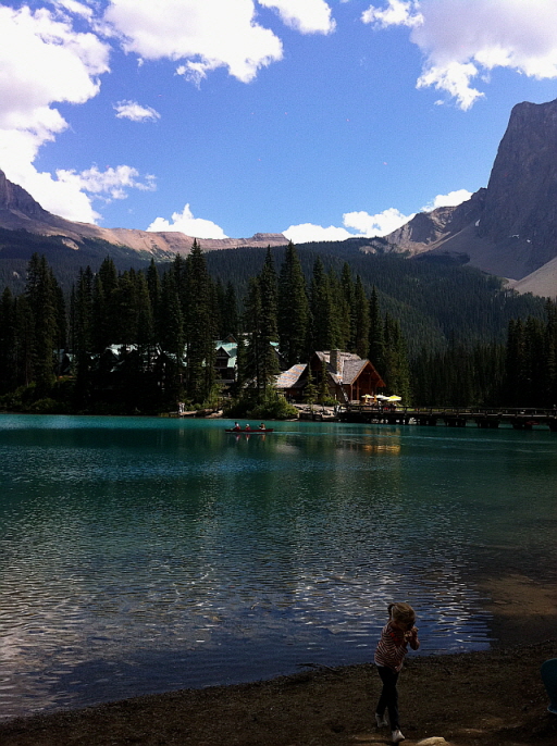 에메랄드빛 호수 앞에 서 있는 아이, 뒤편에는 통나무 롯지들과 숲, 산이 어우러짐.