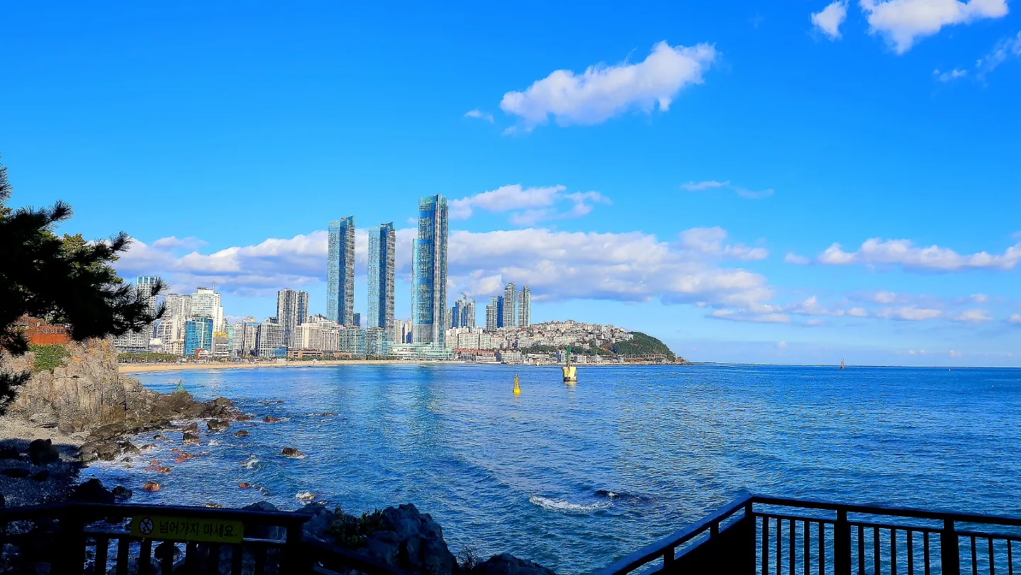 부산 해운대 바다와 광안대교 일대의 전경, 푸른 하늘과 고층 빌딩이 어우러진 모습.