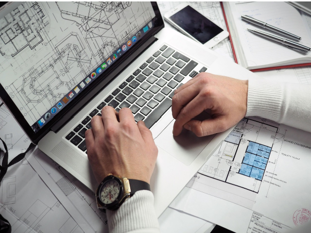 건축 도면 위에서 노트북으로 작업 중인 사람의 손과 설계 자료