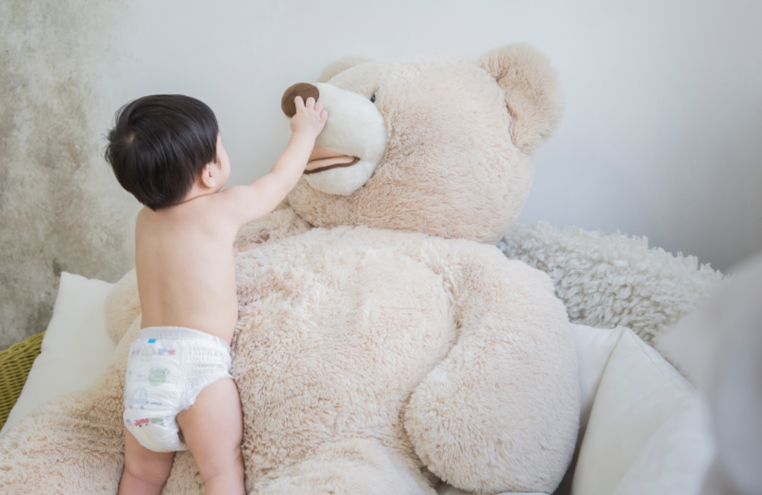 기저귀를 찬 아기가 커다란 곰인형의 코를 만지고 있다.