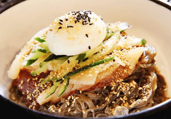 비빔 냉면이 있다. 면 위에 고추장 양념장, 오이채, 배,삶은 달걀 반쪽이 있다.