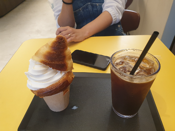 크루아상콘 아이스크림과 아이스 아메리카노가 노란 테이블 위에 올려져 있다. 이 메뉴들을 바라보는 한 사람