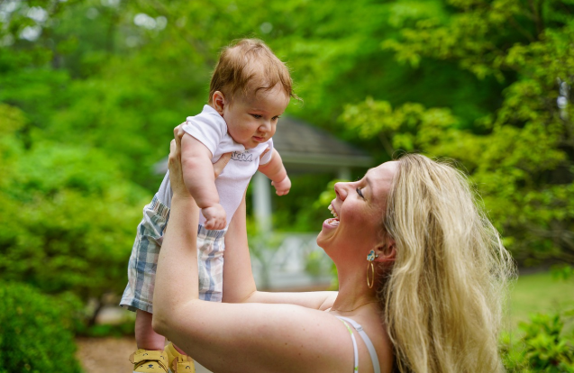 엄마가 아기를 들어 올리며 환하게 웃고 있는 모습. 배경은 초록이 무성한 자연 풍경.