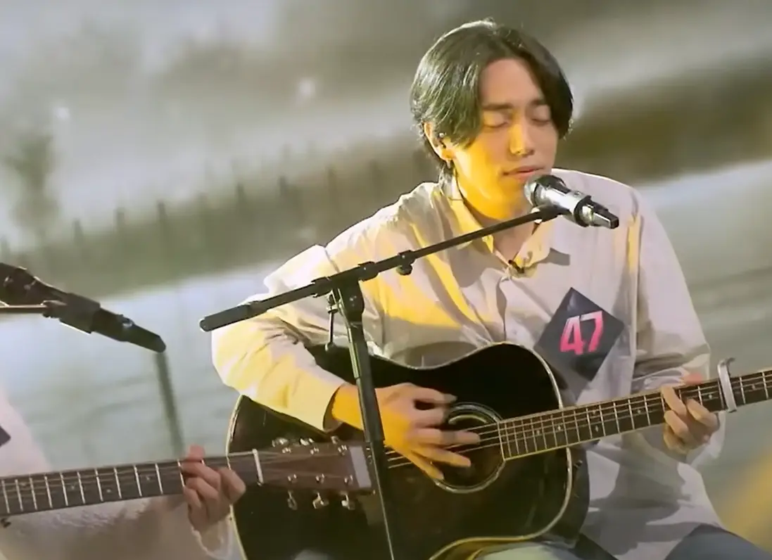 싱어게인3- 베이지색셔츠 가슴에 47번호표를 달고 검은색 기타를 치며 스탠딩마이크를 앞에 두고 두눈 감은채 노래하는 47호 