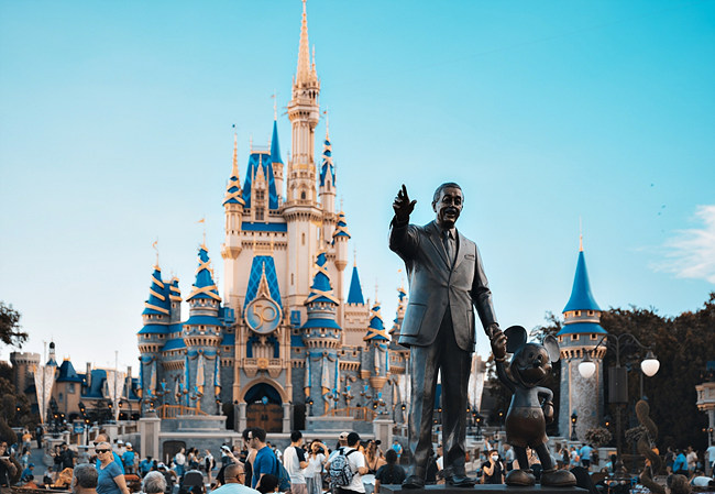 월트-디즈니와-미키-마우스-동상이-앞에-있고-뒤에-아름다운-성이-있으며-그-사이에-놀러온-많은-사람들이-있다